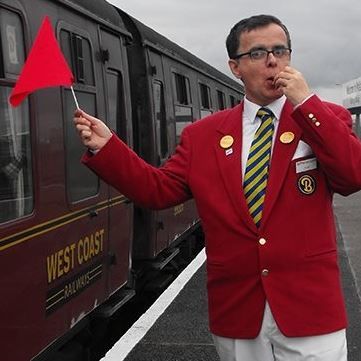 Frankie holds a red flag and blows a whistle on the platform by a steam train