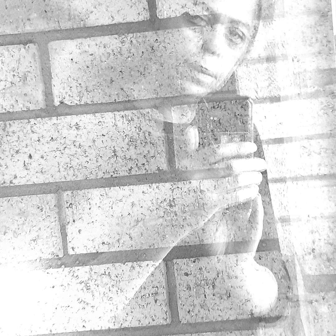 In black and white, Nettie is visible taking a mirror selfie through a semitransparent brick wall.