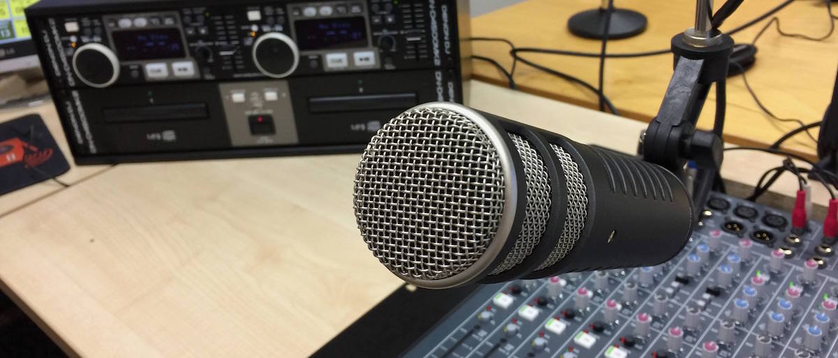 A microphone points towards the camera, with a mixing desk in the background.